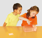 Das Bild zeigt zwei Kinder, die an einem Tisch mit einem transparenten Behälter arbeiten. Das Mädchen in orangefarbenem Shirt untersucht den Behälter, während der Junge in gelbem Shirt interessiert zuschaut.