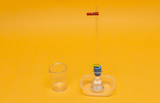 Das Bild zeigt ein Experimentier-Set, das aus einem Behälter mit Flüssigkeit, einer Glasröhre und einem kleinen Messbecher besteht. Alles ist auf einem leuchtend orangefarbenen Hintergrund angeordnet, was die Geräte hervorhebt.
