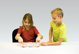Das Bild zeigt zwei Kinder, die an einem Tisch stehen und mit ihren Fingern in drei Gläser mit Flüssigkeit tauchen. Die Gläser sind beschriftet und scheinen Teil eines Experimentes zu sein, das verschiedene Substanzen untersucht.