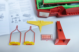 Das Bild zeigt ein Experimentier-Set mit verschiedenen Messinstrumenten, darunter zwei gelbe und rote Halterungen sowie eine Schachtel mit Messkugeln. Dazu liegt ein Handbuch aufgeschlagen, das Anleitungen zur Durchführung von Experimenten enthält.