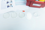 Das Bild zeigt Materialien für ein Experiment, darunter mehrere Petri-Schalen, eine Pipette und einen Messbecher. Zudem ist ein geöffnetes Handbuch sichtbar, das Anweisungen für die Durchführung des Experiments enthält.