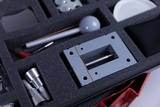 Das Bild zeigt ein aufgeräumtes Set von Experimentiermaterialien, das in einer schaumstoffgepolsterten Box gespeichert ist. Es sind verschiedene wissenschaftliche Geräte und Zubehörteile wie Metallteile und ein grauer Block sichtbar, die für Experimente im Bildungskontext verwendet werden können.
