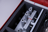 Das Bild zeigt eine rote Box, die mit Schaumstoff ausgekleidet ist und verschiedene experimentelle Geräte enthält. Im Inneren sind mehrere metallische Komponenten mit Schraubnüssen und einer Halterung zu erkennen, die für wissenschaftliche Experimente verwendet werden können.