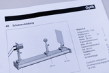 Das Bild zeigt eine Abbildung eines Experimentieraufbaus zur Schattenbildung mit Materialliste. Eine Schiene, zwei Reiter und verschiedene Lichtquellen sind für die Durchführung des Experiments vorgesehen.