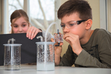 Das Bild zeigt zwei Schüler, die in einer Experimentierumgebung arbeiten. Der Junge fokussiert sich auf ein Gerät, während das Mädchen neugierig einen schwarzen Karton hält, vermutlich um einen Teil des Experiments abzudecken.