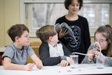 Auf dem Bild sind vier Kinder in einem Klassenzimmer zu sehen, die an einem Experiment arbeiten. Ein Mädchen liest aus einem Buch vor, während ein anderes Kind mit einer Apparatur arbeitet, um eine chemische Reaktion zu beobachten.
