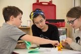 Das Bild zeigt drei Kinder, die gemeinsam an Bastelprojekten arbeiten. Sie benützen farbige Papiere und scheinen engagiert und konzentriert bei der Sache zu sein.