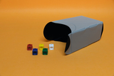 Das Bild zeigt eine graue, gebogene Box mit schwarzer Innenfläche, die auf einem orangefarbenen Hintergrund liegt. Daneben sind mehrere farbige, quadratische Bausteine in rot, gelb, grün, blau und weiß angeordnet.