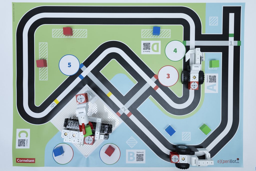 Der Spielplan mit den eXperiBot Robotern zum Thema Recycling ist von oben fotografiert. 
Links gibt es wieder den Grabber, der einen gr&uuml;nen Schaumstoffw&uuml;rfel h&auml;lt. Dieser wird nun auf das Flie&szlig;band gelegt, wo dann durch Drehbewegung entschieden wird, wohin der gr&uuml;ne Schaumstoffw&uuml;rfel abgelegt wird. Rechts sind zwei eXperiBots zu sehen, die sich an markierten Stellen den zu recycelnden M&uuml;ll holen.