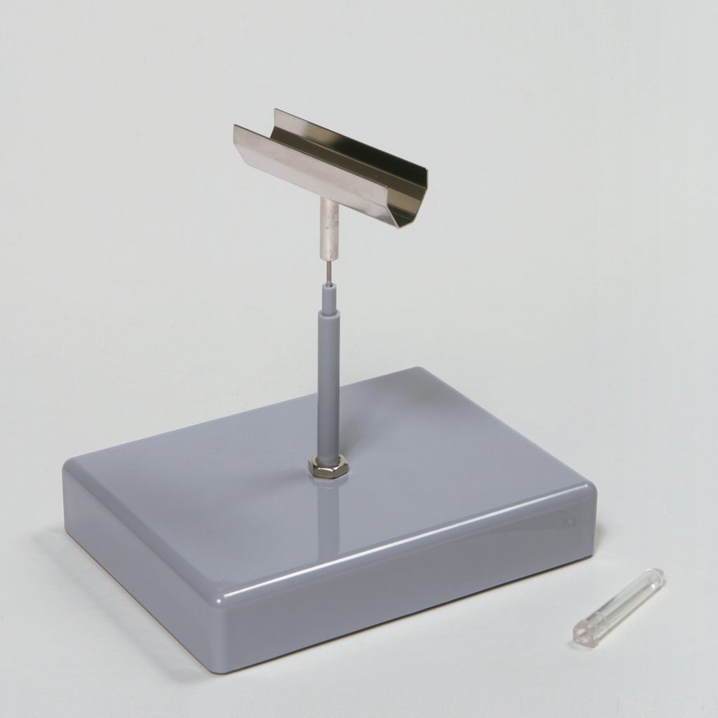 Das Bild zeigt ein Experimentiergerät, bestehend aus einer stabilen grauen Basis mit einem aufrechten Metallarm und einer schmalen Metallplatte oben. Daneben liegt ein durchsichtiges Röhrchen, das wahrscheinlich für Versuche verwendet wird.