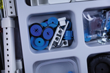 Das Bild zeigt eine Aufbewahrungsbox mit verschiedenen Experimentiermaterialien, darunter blaue und schwarze Scheiben sowie weiße Teile und Verbindungselemente. Diese Materialien sind wahrscheinlich für physikalische Experimente im Bildungsbereich konzipiert.