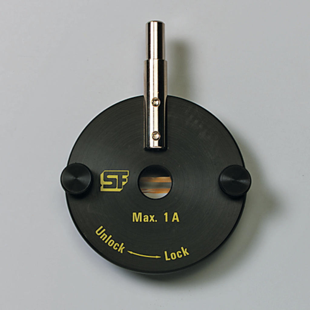 Das Bild zeigt ein schwarzes experimentelles Gerät mit einem Metallständer in der Mitte. Es ist mit der Aufschrift "Max. 1 A" und "Unlock" sowie "Lock" versehen, was auf eine Funktion zur Steuerung von elektrischen Strom hinweist.