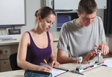 Das Bild zeigt zwei Schüler, die an einem Experimentieraufbau arbeiten, der anscheinend mit Windkraft zu tun hat. Die Schülerin notiert etwas, während der Schüler die elektrischen Komponenten des Modells untersucht.