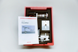 Das Bild zeigt ein Schüler-Set namens "DynaMot", das in einer roten Kunststoffbox präsentiert wird. Neben verschiedenen Bauteilen ist eine Anleitung zu sehen, die die Nutzung des Sets für Experimente im Bildungsbereich beschreibt.