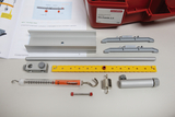 Das Bild zeigt einen Experimentierkasten mit verschiedenen Komponenten, darunter Aluminiumprofile, ein Kraftmessgerät, und Verbindungselemente, die für physikalische Experimente zur Mechanik verwendet werden. Zusätzlich liegt eine Anleitung bei, die die Nutzung der Materialien erklärt.