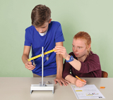 Auf dem Bild sind zwei Schüler zu sehen, die an einem Experiment mit einer Wippe arbeiten. Der Junge hält einen Maßstab, während das Mädchen Notizen auf einem Arbeitsblatt macht, um ihre Ergebnisse festzuhalten.