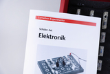 Das Bild zeigt eine Broschüre mit dem Titel "Elektronik" aus einem Schüler-Set von Cornelsen Experimenta. Im Hintergrund sind Komponenten eines Experimentiersets für Elektronik abgebildet, die zur praktischen Erprobung verwendet werden können.