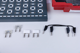 Das Bild zeigt eine Experimentierplatine mit mehreren runden Löchern und eine Reihe von Bauteilen, darunter Widerstände und Kabel mit Klemmen. Diese Komponenten sind typisch für den Einsatz in Experimenten im Lernbereich Elektrotechnik.