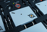 Das Bild zeigt eine Werkzeug- und Experimentierkiste, die verschiedene Geräte und Materialien für schulische Experimente enthält. Dazu gehören graue Platten, Clips, ein Kompass und weitere Utensilien, die in einer Schaumstoffeinlage organisiert sind.