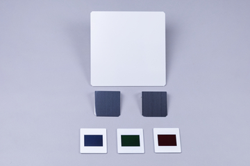 Das Bild zeigt eine Anordnung von Experimentiermaterialien, bestehend aus einer großen weißen Platte und mehreren kleineren farbigen Folien in Blau, Grün und Rot. Neben diesen befinden sich zwei graue quadratische Platten, die wahrscheinlich für wissenschaftliche Experimente verwendet werden.