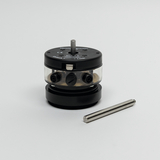 Das Bild zeigt einen Vibrationsgenerator mit einem transparenten Gehäuse und Steuerknöpfen. Neben dem Gerät liegt ein metallischer Stab, der möglicherweise als Zubehör dient.