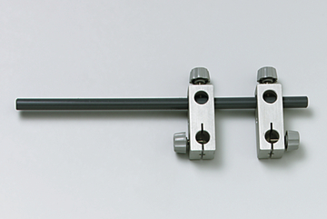 Das Bild zeigt ein Experimentiergerät mit einem grauen Stab und zwei metallischen Halterungen. Diese Halterungen ermöglichen die Befestigung des Stabs in unterschiedlichen Positionen, vermutlich für physikalische Experimente.