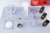 Das Bild zeigt eine Sammlung von Experimentierutensilien, darunter einen Dreibeinständer, Messbehälter, eine Waage, eine Schaufel, und mehrere Glasfläschchen. Diese Materialien sind geeignet für wissenschaftliche Experimente in der schulischen Bildung.