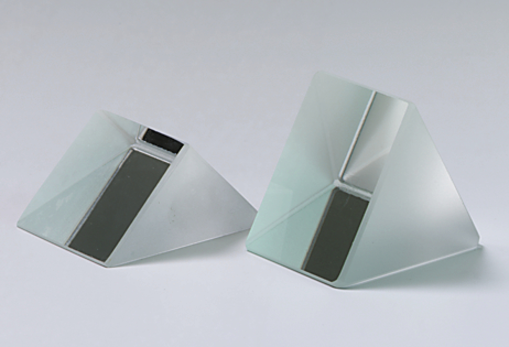 Das Bild zeigt zwei prismatische Glasskörper mit einer matten Oberfläche und einer horizontalen, schwarzen Fläche. Diese geometrischen Objekte werden häufig für physikalische Experimente verwendet, um Lichtbrechung und Reflexion zu demonstrieren.