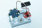 Das Bild zeigt ein elektronisches Experimentierboard mit einer zentralen Platine in Form eines Sterns, die verschiedene elektronische Komponenten enthält. An das Board sind Batteriefach und Steuerungselemente angeschlossen, die zur Durchführung von Experimenten im Bildungsbereich verwendet werden können.