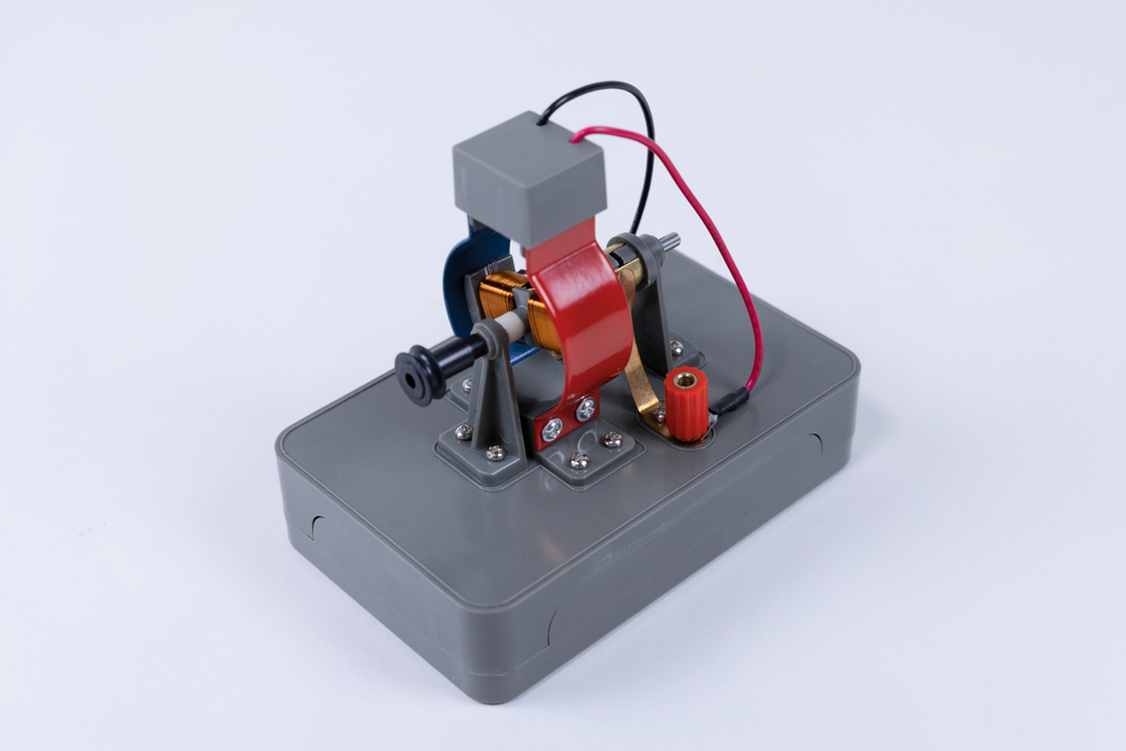 Das Bild zeigt ein graues Experimentiergerät, das aus verschiedenen metallischen Komponenten besteht, darunter eine Spule und elektrische Anschlüsse. Es dient vermutlich der Veranschaulichung grundlegen­der physikalischer Prinzipien, wie Elektromagnetismus oder Motorenbau.