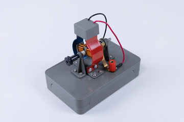 Das Bild zeigt ein graues Experimentiergerät, das aus verschiedenen metallischen Komponenten besteht, darunter eine Spule und elektrische Anschlüsse. Es dient vermutlich der Veranschaulichung grundlegen­der physikalischer Prinzipien, wie Elektromagnetismus oder Motorenbau.