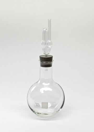Das Bild zeigt ein chemisches Glasgerät, genauer gesagt einen Rundkolben mit einem eingebauten Stopfen und einem Glasrohr. Dieses Laborglasgerät wird häufig in Experimenten zur Durchführung von chemischen Reaktionen verwendet.