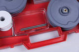 Das Bild zeigt eine rote Aufbewahrungsbox, die verschiedene Experimentiermaterialien enthält, darunter graue Platten, eine Rolle Schnur und ein transparentes Behältnis mit Metallstangen. Diese Utensilien sind wahrscheinlich für schulische Experimente oder Lehrzwecke gedacht.