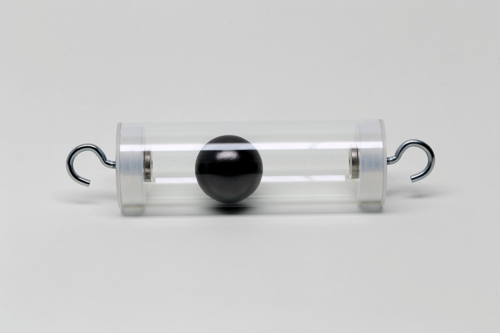Das Bild zeigt einen transparenten Zylinder mit einer schwarzen Kugel in der Mitte, die durch zwei Metallhaken auf beiden Seiten des Zylinders gehalten wird. Dieses Experimentiergerät könnte zur Veranschaulichung physikalischer Prinzipien wie Schwingungen oder Gleichgewicht verwendet werden.