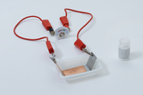 Das Bild zeigt ein Experimentierset mit zwei roten Kabeln, einem Multimeter zur Spannungsmessung und einer kleinen Kunststoffschale, die mit einer Kupfer- und einer Metallplatte ausgestattet ist. Neben dem Set befindet sich ein kleines Glas mit Zitronensäure, das für chemische Experimente verwendet werden kann.