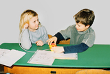 Das Bild zeigt zwei Kinder, die an einem Tisch sitzen und mit einem Experimentiermaterial arbeiten. Das Mädchen beobachtet den Jungen, der ein buntes Gerät in der Hand hat und eine Aufgabe aus einem Arbeitsblatt löst.