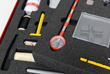 Das Bild zeigt eine Auswahl an Experimentiermaterialien, die in strukturiertem Schaumstoff aufbewahrt sind. Zu sehen sind verschiedene Werkzeuge und Materialien, darunter ein Messgerät, ein Behälter und unterschiedliche Schreib- beziehungsweise Bastelutensilien.