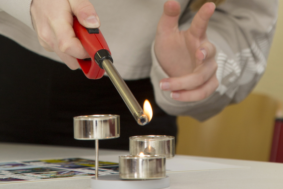 Eine Person verwendet ein Feuerzeug, um eine Kerze anzuz&uuml;nden, w&auml;hrend sie dabei eine Handgeste macht. Auf dem Tisch liegen weitere Teelichter und Materialien f&uuml;r Experimente, die f&uuml;r den Bildungsbereich geeignet sind.