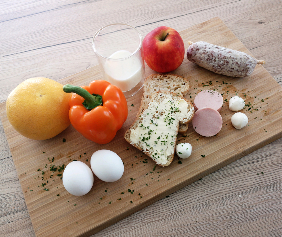 Auf einem Holzbrett sind verschiedene Lebensmittel sortiert: ein gelber Melon, eine orange Paprika, ein Apfel, mehrere Eier, Brot mit Butter, Wurstaufschnitt und Mozzarella-B&auml;llchen. Neben den Lebensmitteln liegen frische Kr&auml;uter verstreut, was das Arrangement appetitlich aussehen l&auml;sst.