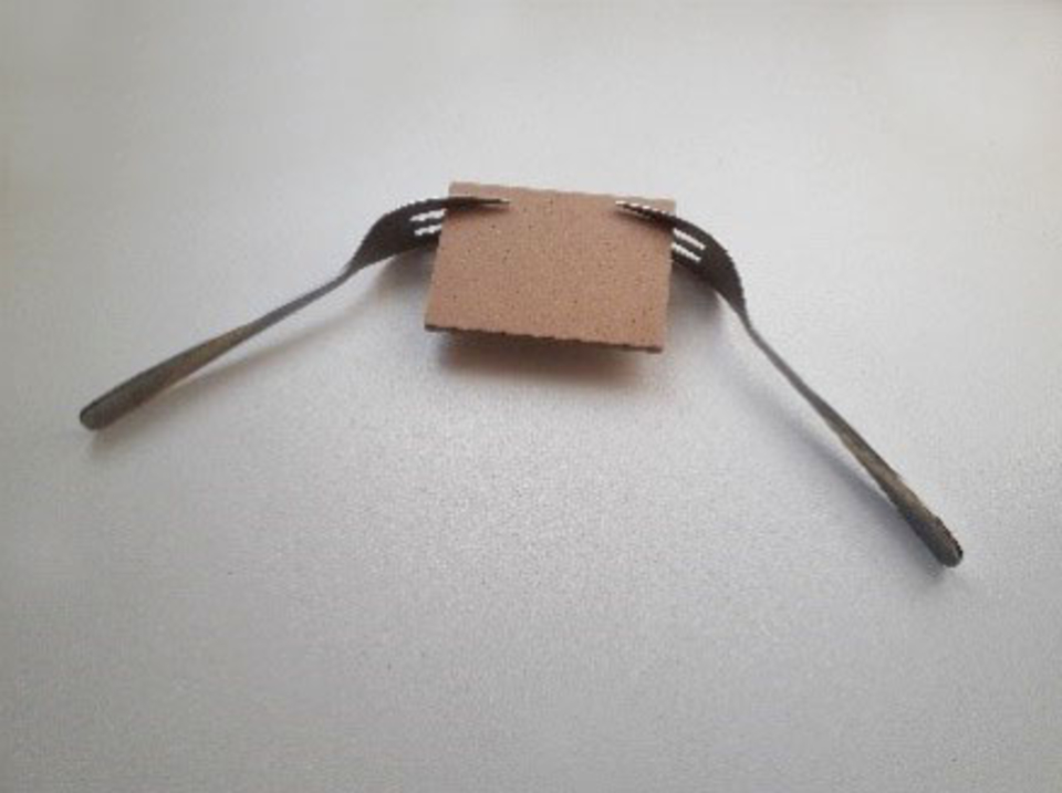 Zwei Gabeln sind an einem kleinen, quadratischen St&uuml;ck Karton befestigt, das in der Mitte des Bildes liegt. Die Gabeln ragen nach au&szlig;en und scheinen eine Art Halterung oder Unterst&uuml;tzung f&uuml;r das Kartonst&uuml;ck zu bilden.