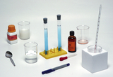 Auf einem weißen Untergrund sind verschiedene Laborgeräte und Chemikalien für Experimente angeordnet. Zu sehen sind Reagenzgläser mit blauer Lösung, Messbecher, Pipette, Löffel und Flaschen mit eindeutig gekennzeichneten Substanzen.