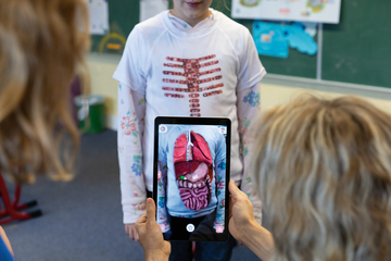 Eine Schülerin trägt ein T-Shirt mit einem Abdruck des menschlichen Körpers, das die inneren Organe zeigt. Jemand hält ein Tablet, das eine digitale Überlagerung der Organe auf dem T-Shirt anzeigt, um den anatomischen Aufbau interaktiv zu erläutern.