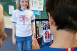 Ein Kind steht in einem Klassenzimmer und trägt ein T-Shirt mit anatomischen Abbildungen. Ein anderes Kind hält ein Tablet, das eine digitale Darstellung des T-Shirts zeigt, wobei Informationen über den Rückenmuskel angezeigt werden.