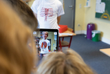 Ein Schüler präsentiert ein T-Shirt mit einem anatomischen Motiv, während ein anderes Kind mit einem Tablet ein Foto davon macht. Im Hintergrund sind Schulmöbel und Lernmaterialien sichtbar, was auf eine Unterrichtssituation hindeutet.