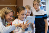 Eine Lehrkraft und mehrere Kinder schauen gemeinsam auf ein Tablet. Die Szene vermittelt Begeisterung für das Lernen und die Nutzung digitaler Medien im Unterricht.