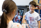Ein Mädchen hält ein Tablet hoch, auf dessen Bildschirm ein Junge mit einem T-Shirt zu sehen ist, das eine anatomische Darstellung zeigt. Der Junge lächelt, während sich die beiden in einem Klassenzimmer mit Lernmaterialien im Hintergrund befinden.