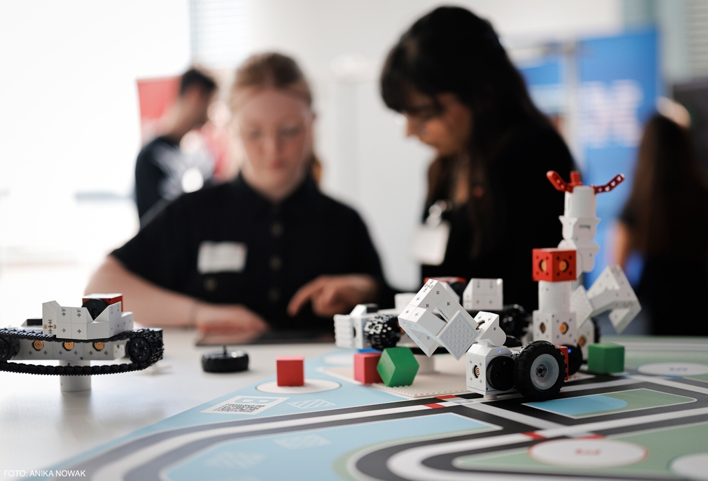 Auf dem Tisch sind verschiedene Lernroboter auf einer Matte platziert., Im Hintergrund sind junge Frauen zu sehen, die sich mit Programmieren der Lernrobotern besch&auml;ftigen und an einem Lernprojekt arbeiten.