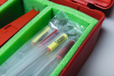 Im roten Kasten sind verschiedene Experimentierutensilien untergebracht, die in grüner Schaumstoffpolsterung stabilisiert sind. Zu sehen sind mehrere durchsichtige Röhrchen mit farbigen Markierungen, die für Bildungsexperimente verwendet werden.