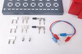 Eine Sammlung von Experimentierkomponenten liegt auf einem weißen Hintergrund. Zu sehen sind verschiedene Steckverbindungen, Sicherungen und Verbindungskabel, die zur Durchführung von elektrischen Experimenten verwendet werden können.