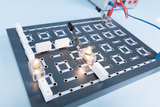 Auf der Platte sind verschiedene Verbindungsstücke und Drähte angeordnet, um elektrische Schaltkreise zu erstellen. Zwei Glühlampen sind installiert und zeigen an, dass der Strom fließt, während die gesamte Anordnung als Experimentierhilfe dient.